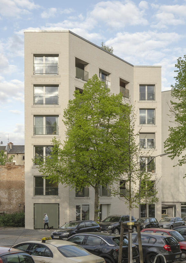 Straßenansicht eines mehrgeschossigen Wohn- und Bürogebäudes mit heller Backstein-Fassade und Loggien; im Vordergrund Bäume und parkende Autos säumen die Straße.